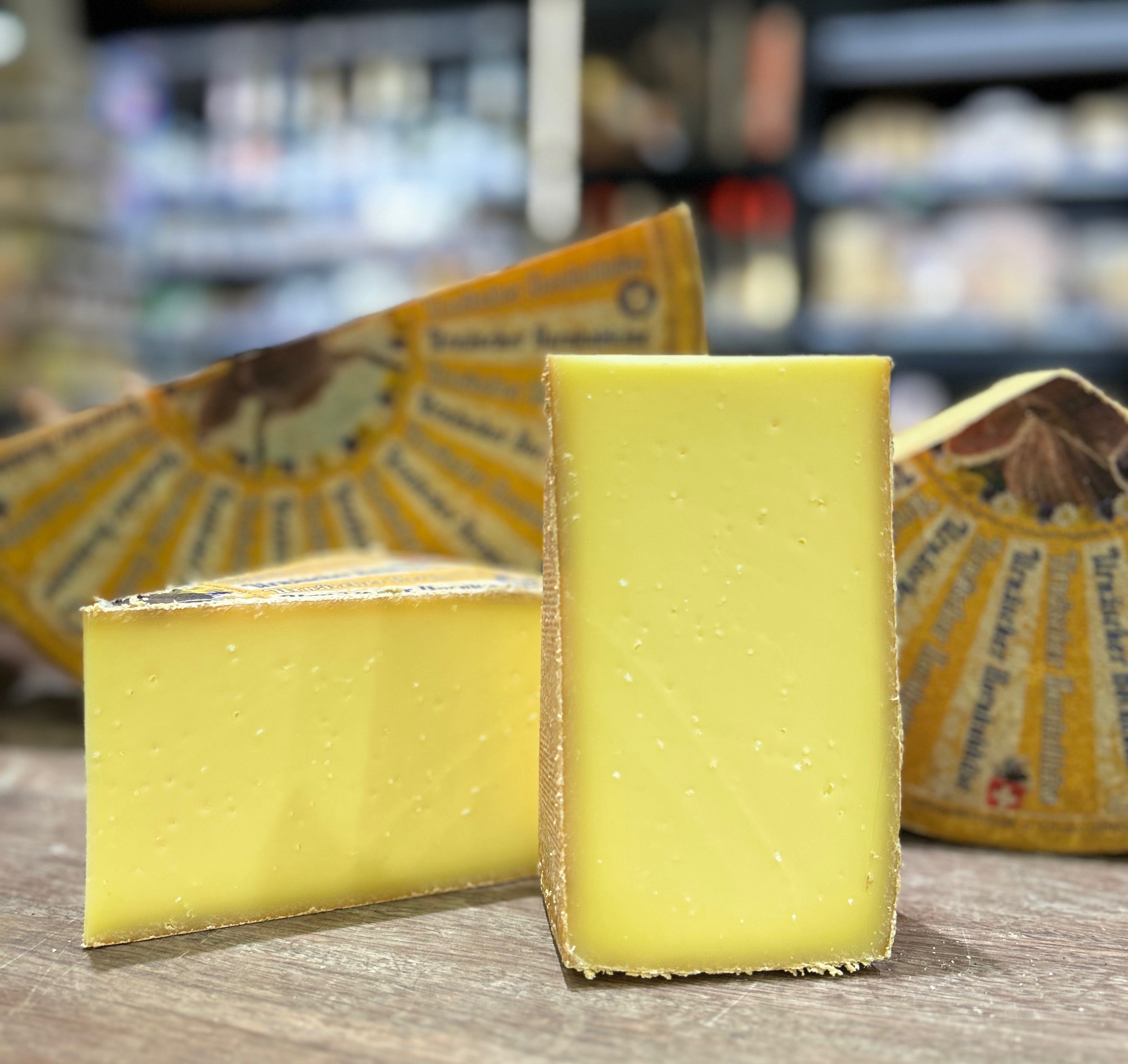 Two wedges of pale yellow cheese standing on a wooden surface, with wrapped cheese blocks in the blurred background of what appears to be a cheese shop.