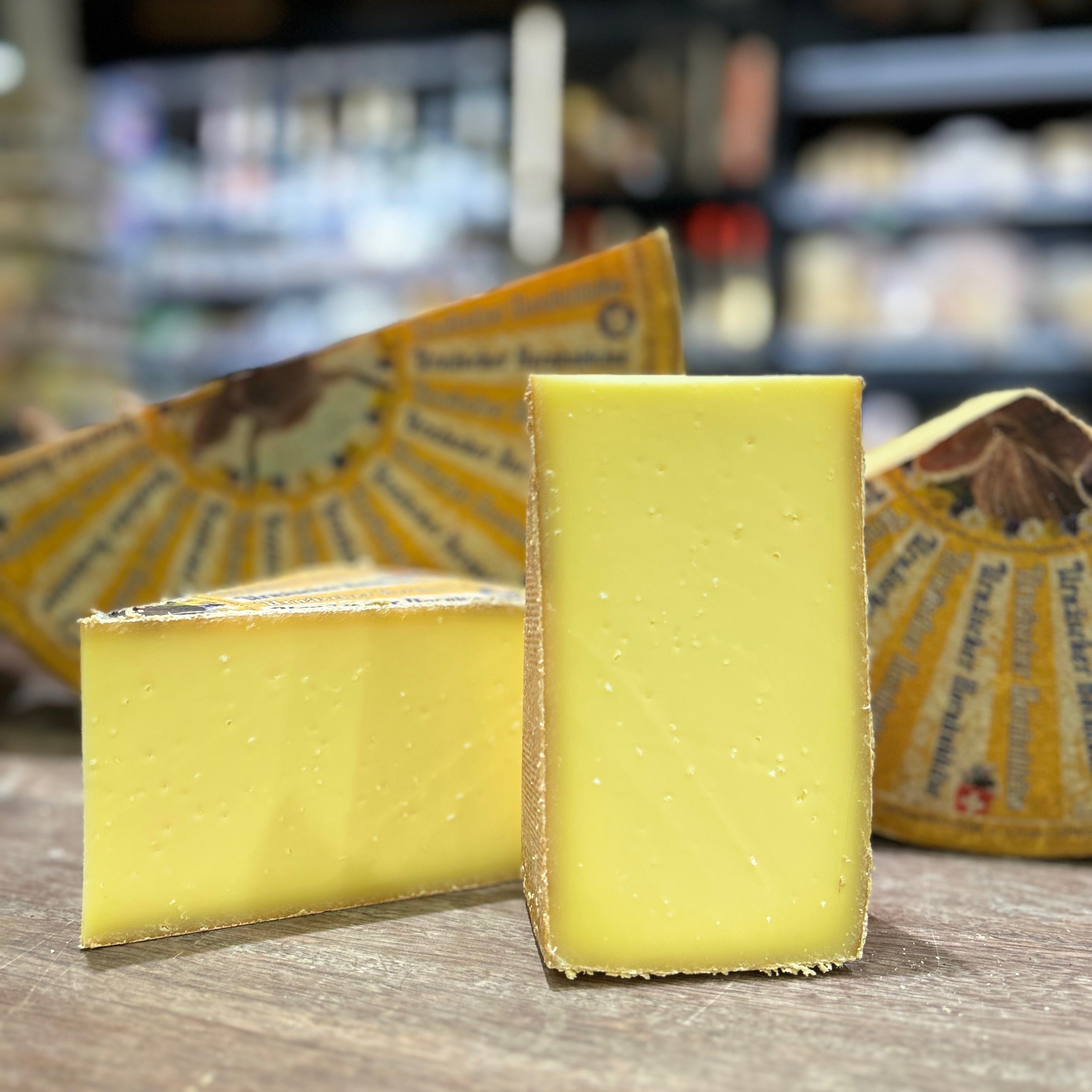 Two wedges of pale yellow cheese standing on a wooden surface, with wrapped cheese blocks in the blurred background of what appears to be a cheese shop.