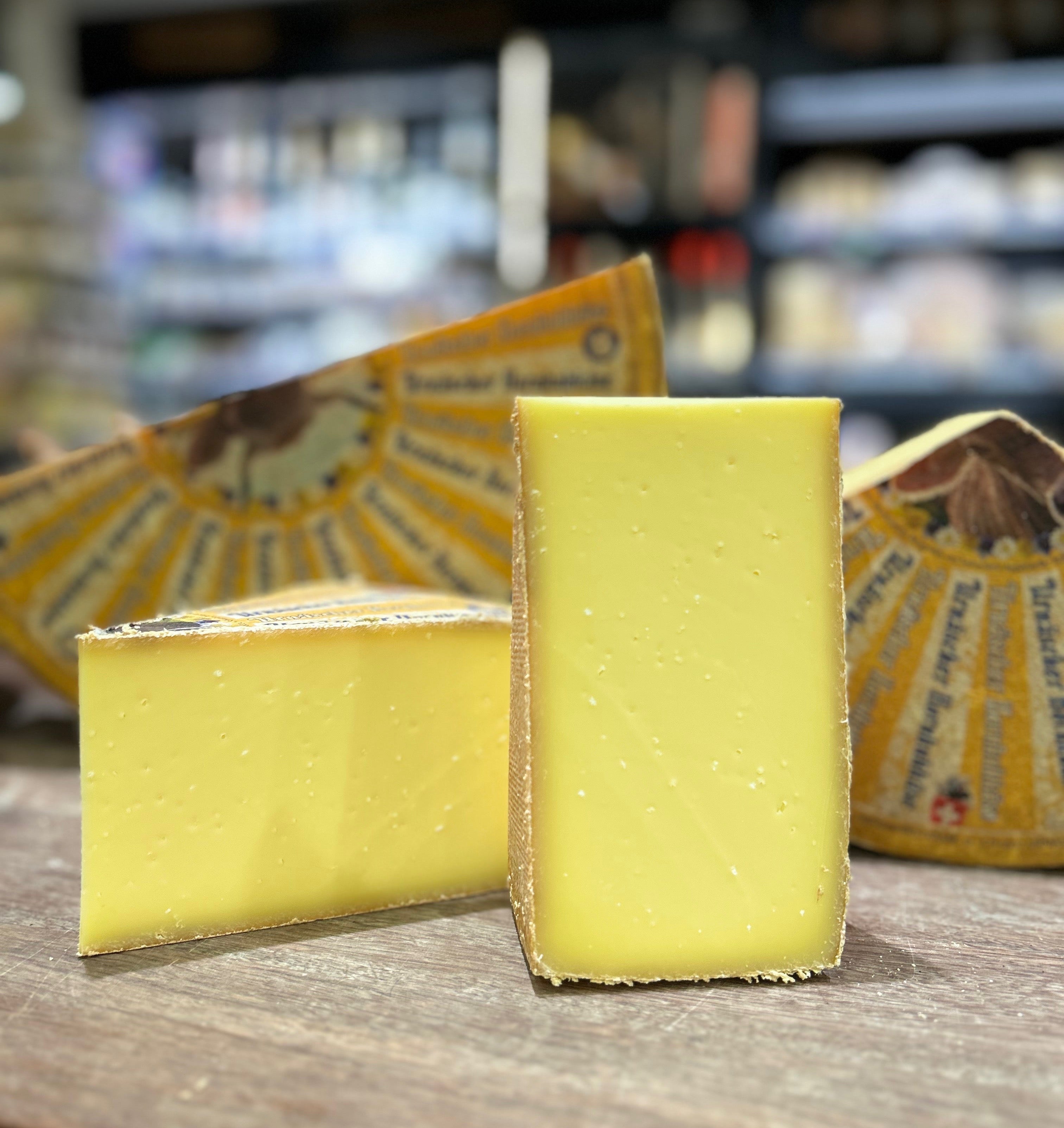 Two wedges of pale yellow cheese standing on a wooden surface, with wrapped cheese blocks in the blurred background of what appears to be a cheese shop.